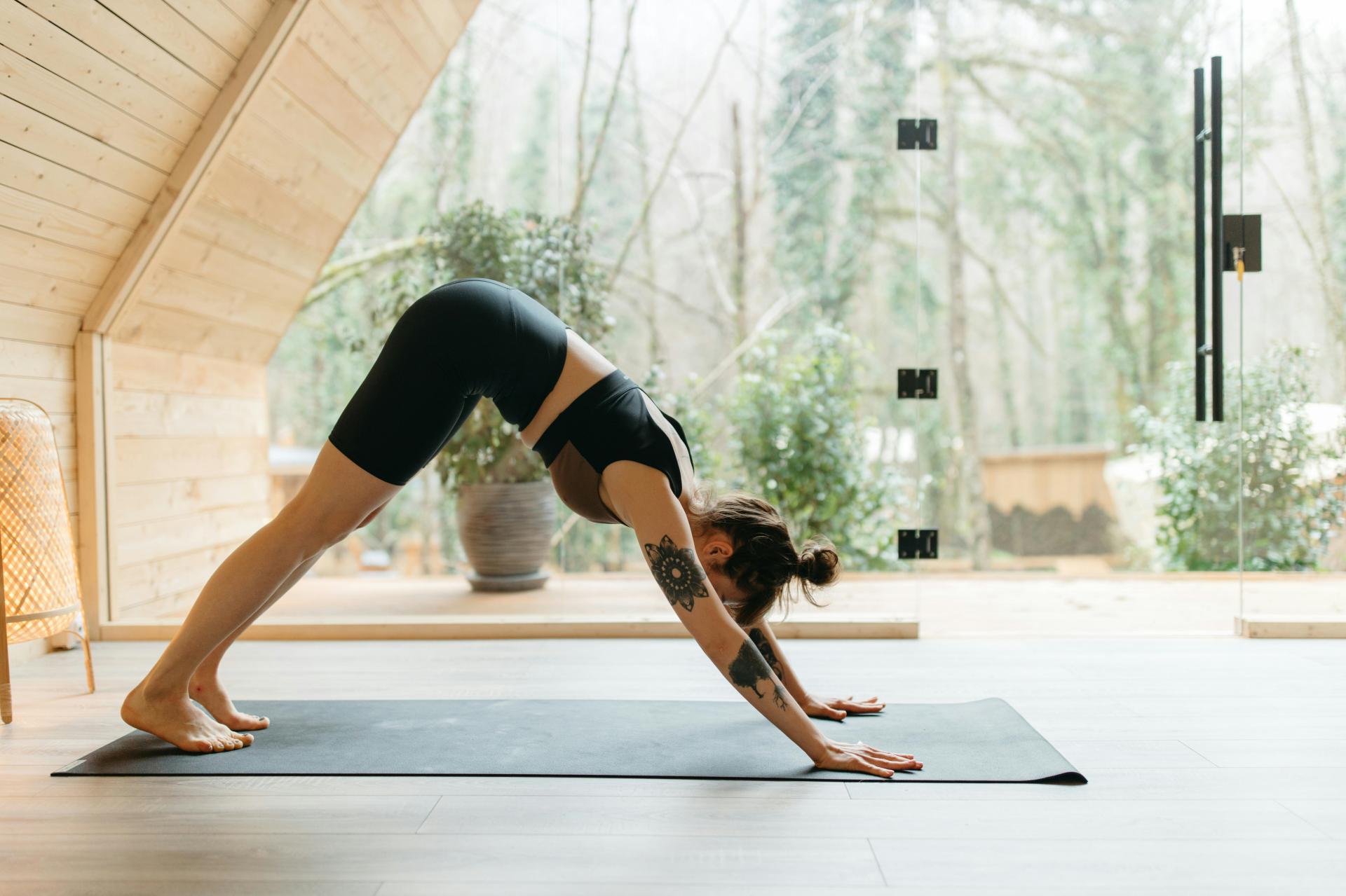 A woman engaged in a Pilates workout, performing a downward dog pose on a yoga mat inside a wooden studio with panoramic views of a forest