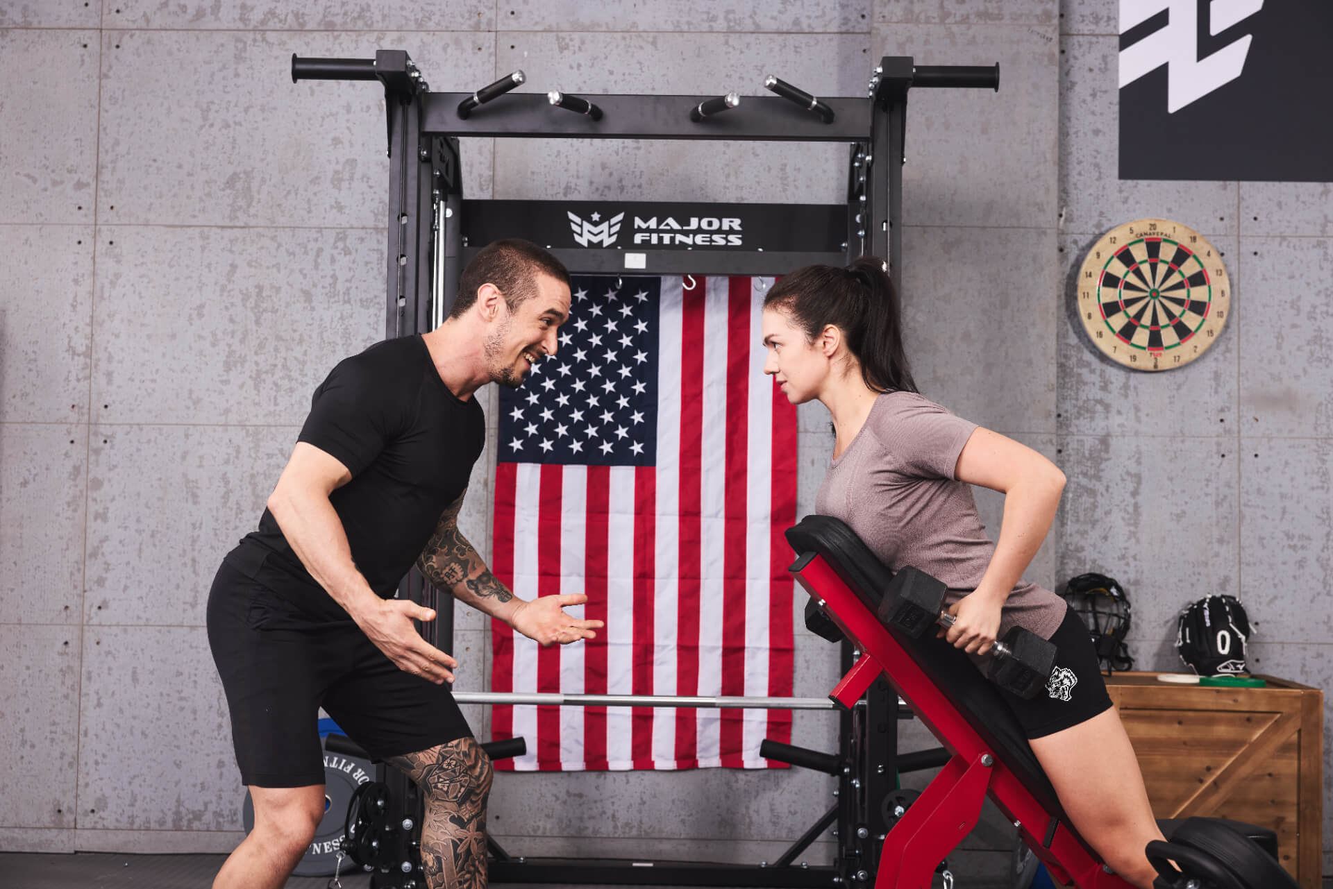  A fitness instructor providing guidance to a female client performing a dumbbell row exercise on a red bench at a Major Fitness home gym