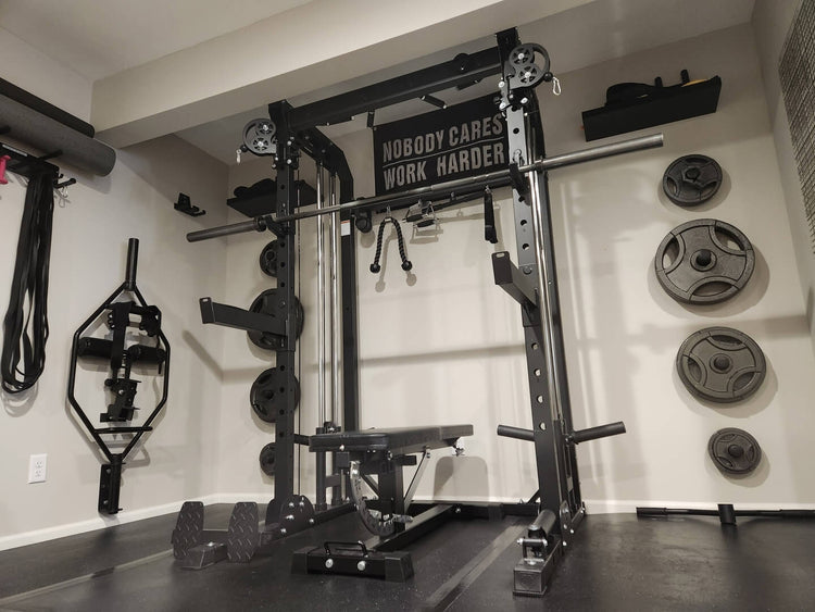 home gym setup with a Smith machine, barbell, weight plates, and a motivational quote on the wall, alongside resistance bands and a hex bar